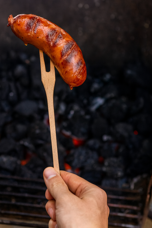 Drewniany widelec grillowy 27 cm z kiełbasą nad grillem – naturalne akcesorium do grillowania, idealne do podawania i obracania mięs.