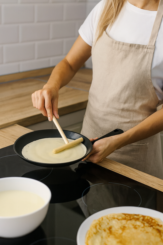 Kobieta w kuchni rozprowadza ciasto naleśnikowe po patelni przy użyciu małego drewnianego rozprowadzacza o długości 20 cm i szerokości części roboczej 14 cm. Obok znajdują się miska z ciastem i talerz z gotowymi naleśnikami. Naturalne oświetlenie podkreśla ciepłe kolory drewna i kuchenne detale. Produkt marki Chłopcy z Rancza