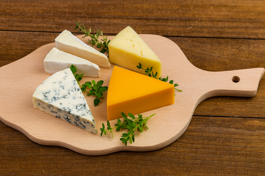 Drewniana deska kuchenna w kształcie liścia z różnymi gatunkami sera – Brie, Cheddar, Emmental i ser pleśniowy – udekorowanymi świeżymi ziołami, ustawiona na rustykalnym drewnianym stole.