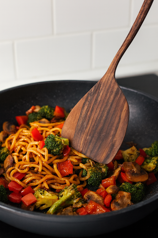 Drewniana łopatka kucharska z ciemnego orzecha miesza warzywa stir-fry na patelni – makaron, brokuły, papryka i pieczarki w jasnej kuchni z białymi płytkami w tle. Styl e-commerce, ciepłe oświetlenie i naturalne drewno.