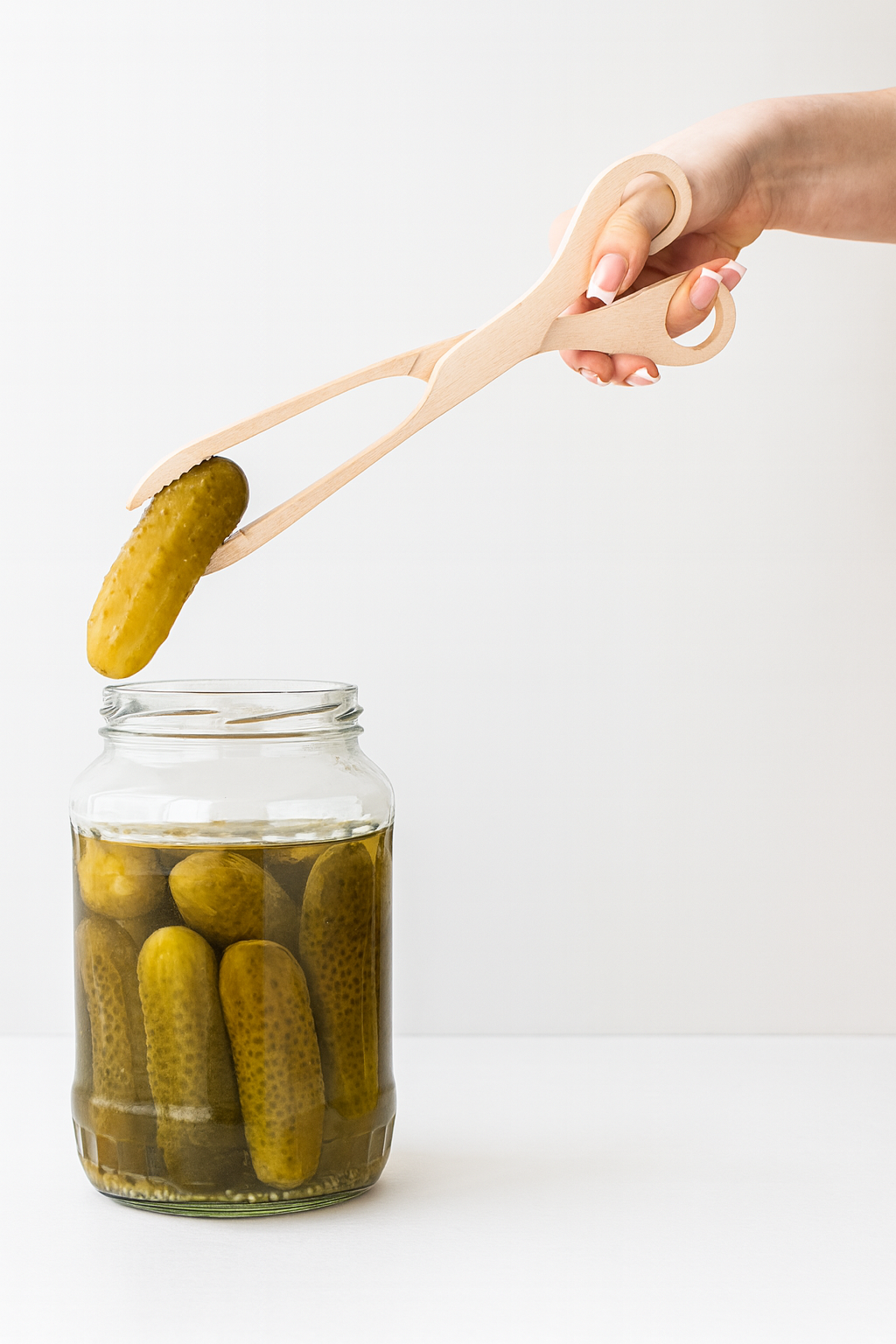 Drewniane szczypce kuchenne chwytające ogórka kiszonego wyjmowanego ze słoika wypełnionego innymi ogórkami w zalewie.