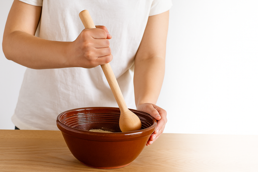 Kobieta wyrabia rzadkie ciasto w brązowej makutrze, używając drewnianej pałki z kulistą końcówką. Drewniane narzędzie ma długość około 30 cm, a misa stoi na jasnym blacie kuchennym w naturalnym oświetleniu.