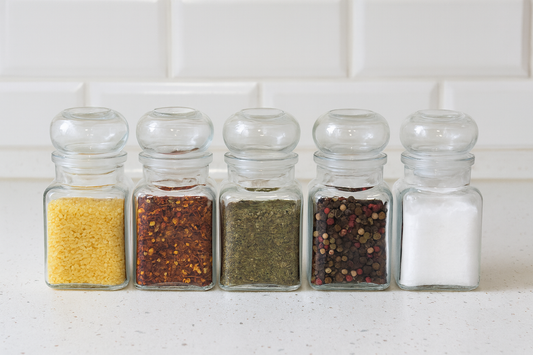 Zestaw pięciu eleganckich szklanych słoików o pojemności 150 ml, ustawionych w rzędzie na kuchennym blacie. Wypełnione są kolejno: drobnym makaronem, suszoną papryką, ziołami, kolorowym pieprzem i solą. Idealne do przechowywania przypraw i dodatków w stylowej kuchni.