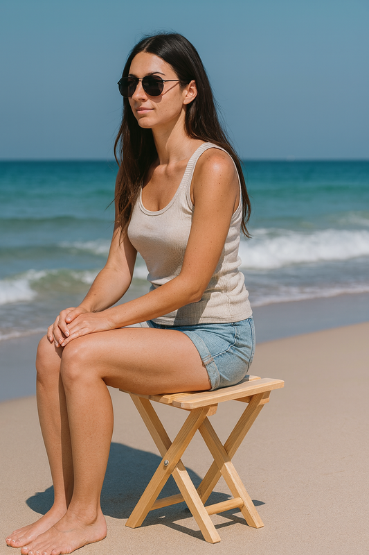 Drewniane składane krzesełko ogrodowe z naturalnego buku, na którym siedzi brunetka w okularach przeciwsłonecznych na plaży. W tle widać morze i jasny piasek