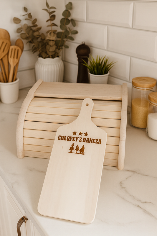 Drewniany chlebak kuchenny z naturalnego drewna bukowego z deską do krojenia z grawerem „Chłopcy z Rancza”, ustawiony na blacie jasnej kuchni w rustykalnej aranżacji — obok słoiki, rośliny i przybory kuchenne.