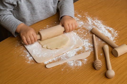 Dziecko rozwałkowuje ciasto wałkiem na małej drewnianej stolnicy o wymiarach 23,5 × 16 cm. Wokół rozsypana mąka i leżą drewniane przybory kuchenne – łyżka, młotek, miodownik i nożyk. Naturalne światło, ciepły, realistyczny klimat kuchenny.
