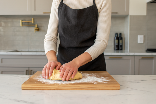 Kobieta wyrabia ciasto na dębowej desce kuchennej w eleganckiej, jasnej kuchni. Deska lekko oprószona mąką leży na marmurowym blacie, a w tle widać nowoczesne szafki i złote uchwyty. | Chłopcy z Rancza