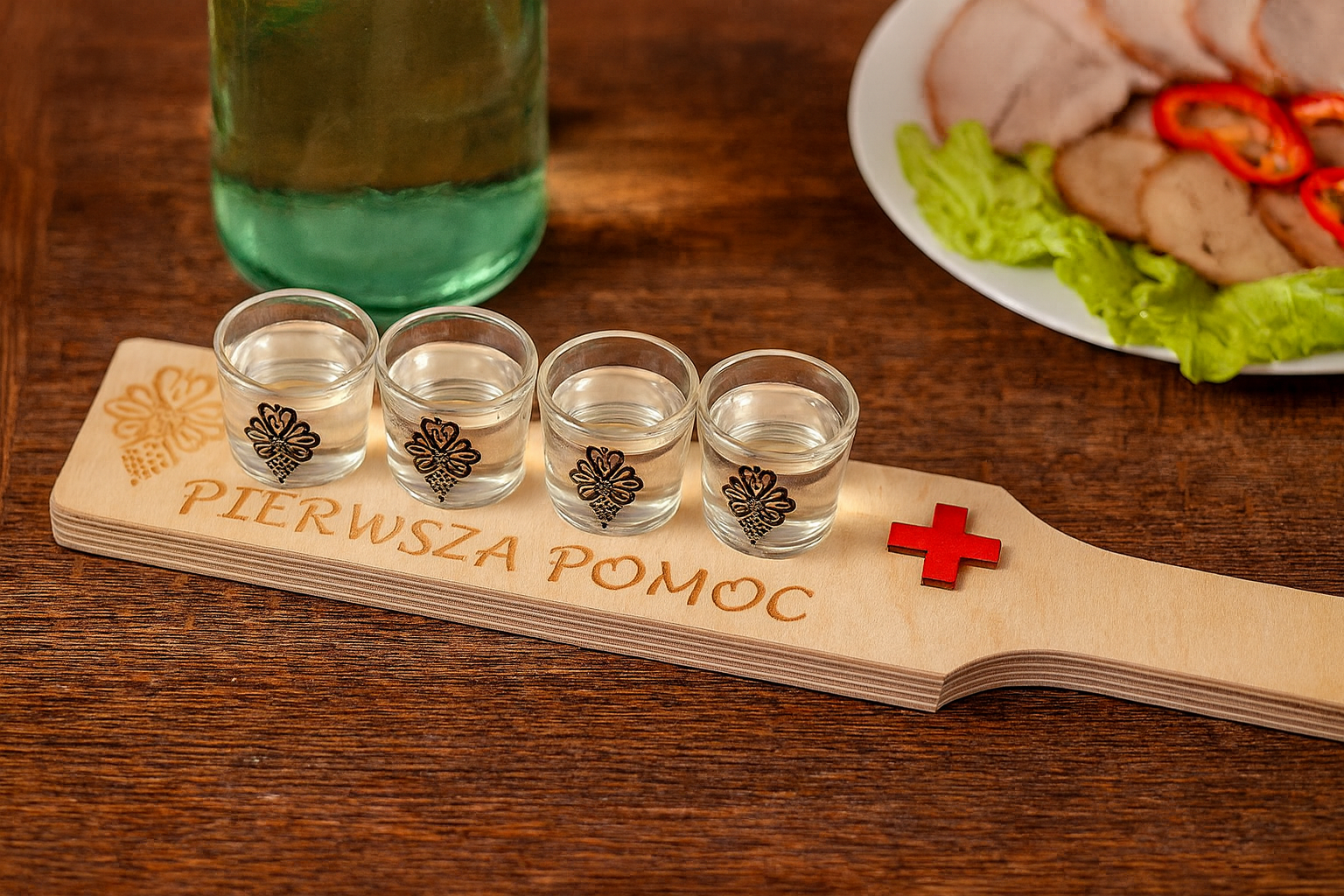 Drewniana taca z napisem Pierwsza Pomoc i czerwonym krzyżem, na której stoją cztery kieliszki napełnione wódką. W tle widać biesiadny stół z butelką i talerzem z wędlinami oraz sałatą, całość w ciepłym, przyjaznym oświetleniu