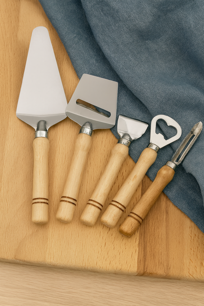 Zestaw czterech stalowych narzędzi kuchennych z drewnianymi rączkami leży na jasnym, drewnianym stole, częściowo przykrytym niebieską lnianą ściereczką. W zestawie znajdują się: łopatka do ciasta, krajalnica do sera, otwieracz do butelek i obieraczka do warzyw. Całość utrzymana w ciepłej, naturalnej tonacji, idealna do sklepu e-commerce z akcesoriami kuchennymi.
