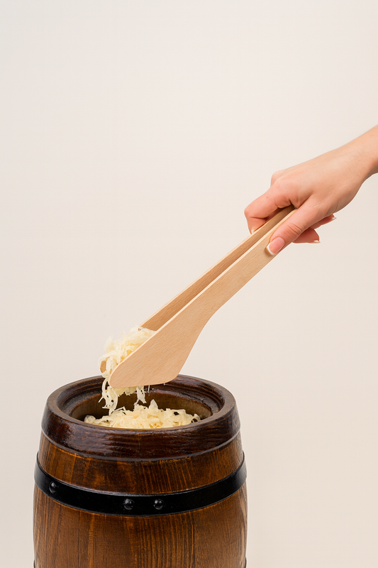 Drewniane szczypce kuchenne wyciągające kiszoną kapustę z tradycyjnej drewnianej beczki – naturalne akcesorium kuchenne w rustykalnym stylu.