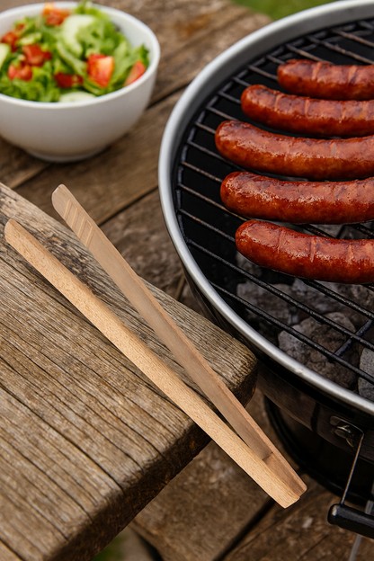 Drewniane szczypce kuchenne leżą na stole obok grilla, na którym grillują się apetyczne kiełbaski. W tle widać miskę z sałatką – naturalna, letnia sceneria idealna do prezentacji akcesoriów grillowych.