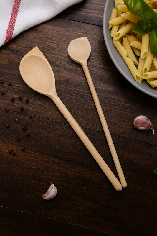 Zestaw dwóch drewnianych łyżek kuchennych leżących na ciemnym stole obok talerza z makaronem, ząbków czosnku i ziaren pieprzu – naturalna sceneria gotowania w kuchni.