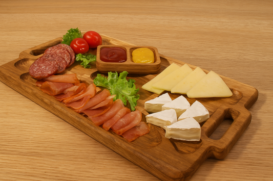 Drewniana deska do serwowania z kilkoma przegródkami, wypełniona różnymi serami, wędlinami oraz świeżą sałatą i pomidorami koktajlowymi. W centralnej miseczce znajdują się dwa sosy – ketchup i musztarda. Całość ułożona na drewnianym stole, w ciepłym świetle, gotowa do podania jako elegancka przekąska.