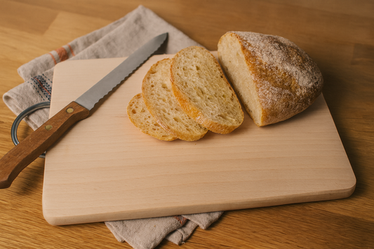 Prostokątna deska bukowa z metalowym uchwytem o wymiarach 20×30 cm i grubości 1,5 cm. Na desce leżą kromki świeżo pokrojonego chleba i nóż z drewnianą rękojeścią. Pod deską lniany ręcznik, tło z jasnego drewna, naturalne ciepłe światło w domowej kuchni.