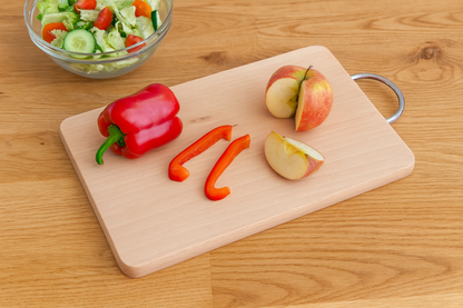 Deska bukowa z metalowym uchwytem o wymiarach 22×33 cm i grubości 1,5 cm. Na desce leżą kawałki czerwonej papryki i pokrojone jabłko. Obok znajduje się miska z sałatką z warzyw – ogórkiem, pomidorkami i sałatą. Tło stanowi blat z naturalnego drewna, oświetlony ciepłym, dziennym światłem.