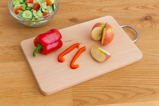 Deska bukowa z metalowym uchwytem o wymiarach 22×33 cm i grubości 1,5 cm. Na desce leżą kawałki czerwonej papryki i pokrojone jabłko. Obok znajduje się miska z sałatką z warzyw – ogórkiem, pomidorkami i sałatą. Tło stanowi blat z naturalnego drewna, oświetlony ciepłym, dziennym światłem.