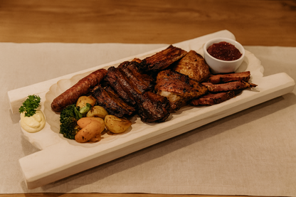 Duże koryto drewniane o wymiarach 35×80 cm, wypełnione biesiadnym zestawem potraw: żeberka, kiełbasa, boczek, pieczony drób, ziemniaki, warzywa i sos w miseczce. Scena w karczmie na drewnianym stole z lnianym obrusem, ciepłe światło, rustykalny klimat.