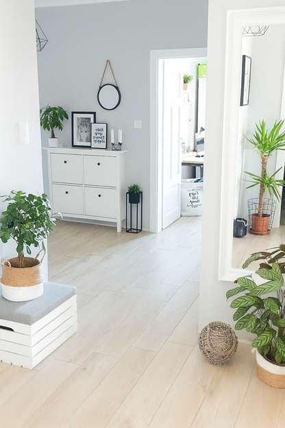 Jasne i nowoczesne wnętrze w stylu minimalistycznym – korytarz z jasną drewnianą podłogą, białymi ścianami i meblami. Widać białą komodę z dekoracjami, lustro w ramie oraz kilka zielonych roślin doniczkowych w plecionych osłonkach. Naturalne światło podkreśla czystość i przytulny charakter pomieszczenia.