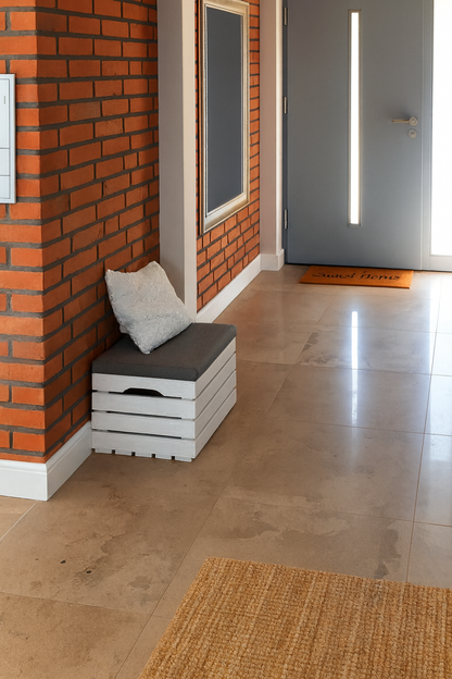 Nowoczesny przedpokój z ceglaną ścianą, jasną podłogą z płytek i szarą ławką wykonaną z białej drewnianej skrzyni z poduszką. W tle widoczne szare drzwi wejściowe i wycieraczka, całość w ciepłym świetle dziennym.